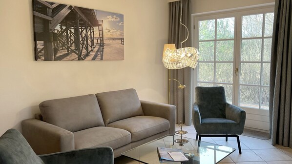 A living room with a sofa, armchair, and a glass coffee table. There's a large window with curtains, a floor lamp, and a painting on the wall.