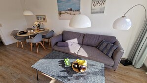 A modern living room with a grey sofa, a dining table with chairs, and a coffee table with fruits and a glass of water. - Luna31 (Room category) - Luna IV (Sankt Peter-Ording)