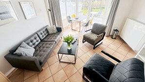 Un salon moderne comprenant un canapé, un fauteuil et une table basse, un coin repas avec des chaises, et une vue dégagée grâce à de grandes fenêtres.