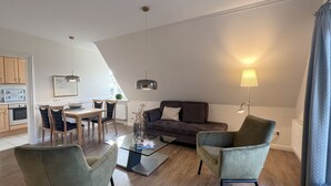 Ein modernes Wohnzimmer mit einem Glastisch, einer Couch und zwei Sesseln. Es gibt einen Essbereich mit Tisch und Stühlen sowie eine Küche mit Schränken und einer Mikrowelle.