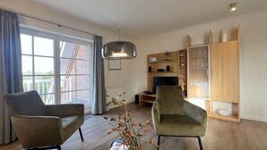 Ein modernes Wohnzimmer mit zwei Sesseln, einem Fernseher, einer Holzkommode und einem großen Fenster mit Blick auf ein Backsteinhaus.