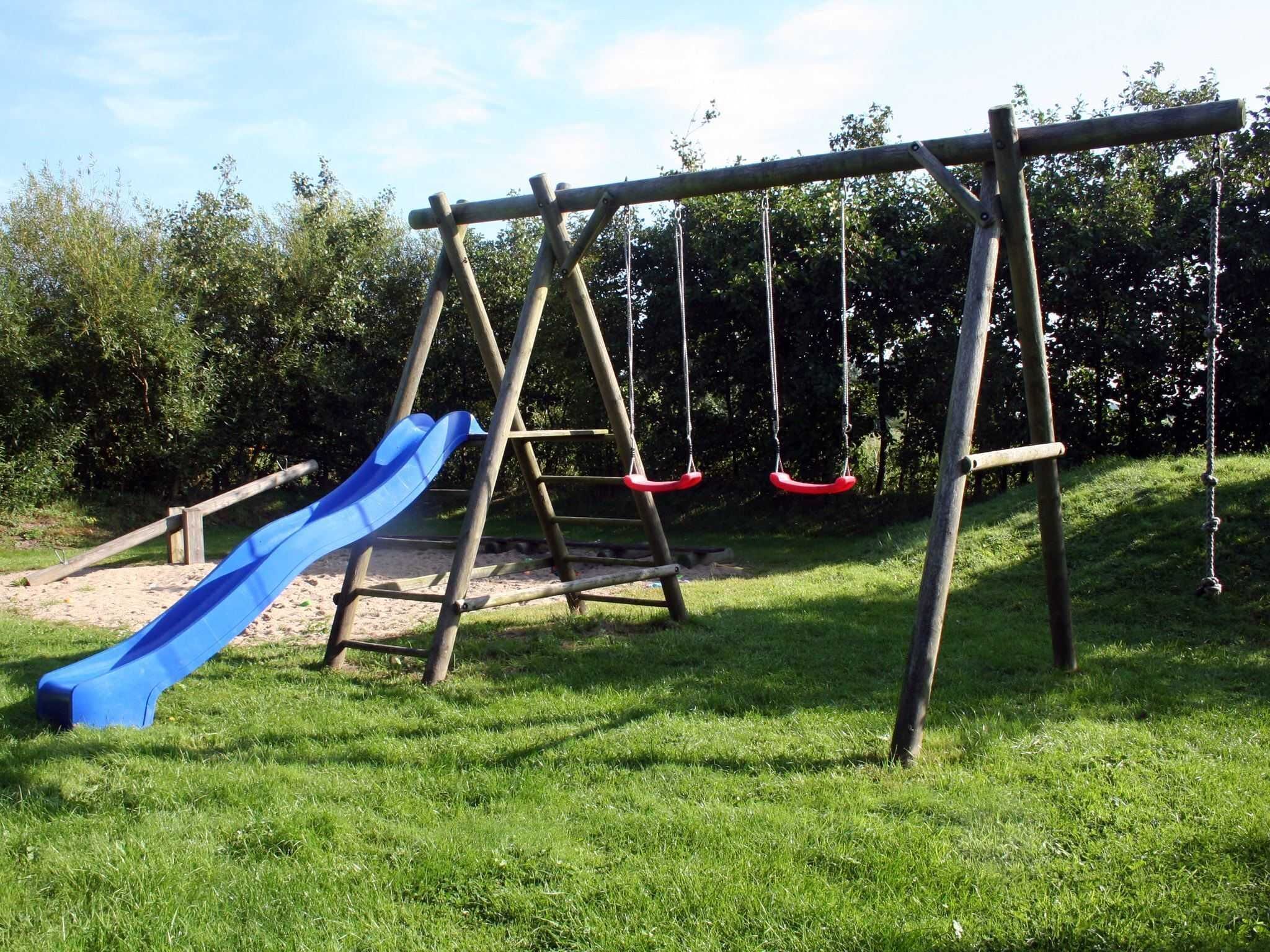 Une aire de jeux avec un toboggan bleu, des balançoires et une structure de balançoire en bois.