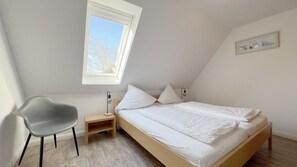 A bedroom with a sloped ceiling, a bed with white bedding, a gray chair, a bedside table, and a small shelf.