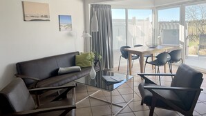 A living room with a sofa, two armchairs, a glass coffee table, and a dining table with chairs. There is a window with curtains, a lamp, and two framed pictures on the wall. - Luna18 (Room category) - Luna II (Sankt Peter-Ording)