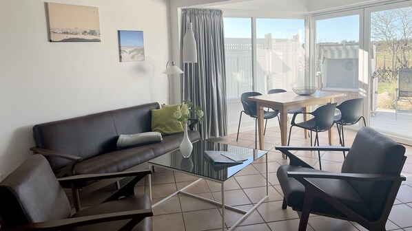 A living room with a sofa, two armchairs, a glass coffee table, and a dining table with chairs. There is a window with curtains, a lamp, and two framed pictures on the wall.