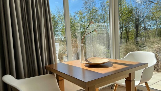 Uma sala de jantar moderna com mesa de madeira e cadeiras brancas, um vaso com flores e vista para árvores e céu através de grandes janelas.