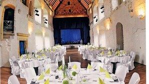 Salão de festas com mesas redondas dispostas para um evento formal, com toalhas de mesa brancas e cadeiras, um palco na frente e paredes de pedra.