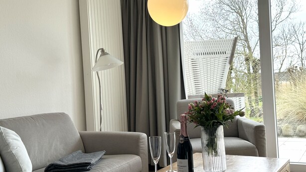 A modern living room with a sofa, a table with a vase of flowers, two wine glasses, and a bottle of wine. - Luna21 (room category) - Luna III (Sankt Peter-Ording)