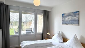 A hotel room with a large window, a bed with white linens, a nightstand with a lamp, and a painting on the wall. - Luna21 (room category) - Luna III (Sankt Peter-Ording)