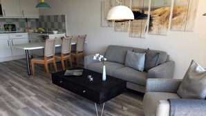 A modern living room with a grey sofa, a black coffee table, and a dining area with wooden chairs and a white table. - Luna21 (room category) - Luna III (Sankt Peter-Ording)