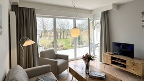 A modern living room with a sofa, a coffee table, a TV, and a large window offering a view of a garden. - Luna21 (room category) - Luna III (Sankt Peter-Ording)