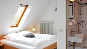 Una camera da letto con un lucernario, un letto con biancheria bianca, una lampada da comodino, un termosifone e un bagno con lavandino e doccia.