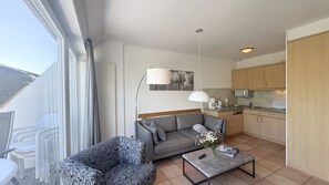 Ein modernes Wohnzimmer mit grauem Sofa, einem gemusterten Sessel und einem Couchtisch mit einer Vase. Im Hintergrund ist eine Küche mit Holzschränken und einer Mikrowelle erkennbar.