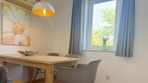Ein Essbereich mit einem Holztisch, grauen Stühlen, einer verzierten Metallschale, einem Daisy-Gemälde und einem Fenster mit blauen Vorhängen.