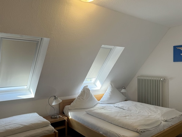 Un dormitorio con techo inclinado, dos ventanas, un radiador y una mesita de noche con lámpara.
