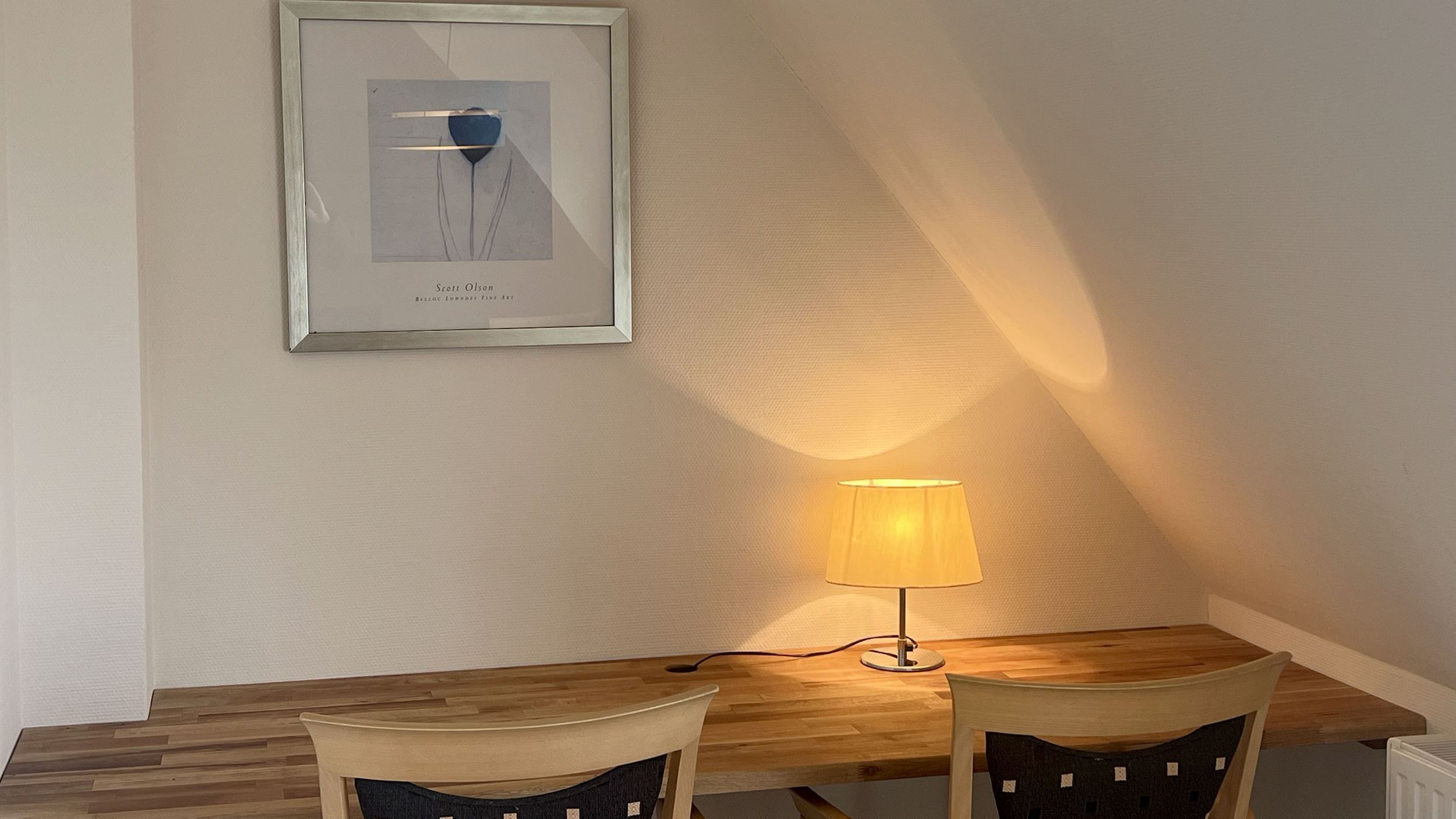 A dining area with a wooden table, two chairs, a framed artwork, and a table lamp.