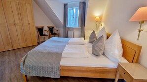 A neatly made bed with pillows and a blanket, a wooden wardrobe, two chairs, a window with curtains, and a wall-mounted lamp.