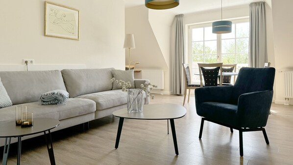 A modern living room with a grey sofa, a dark blue armchair, and a coffee table. There is a window with curtains, a lamp, and a framed picture on the wall.