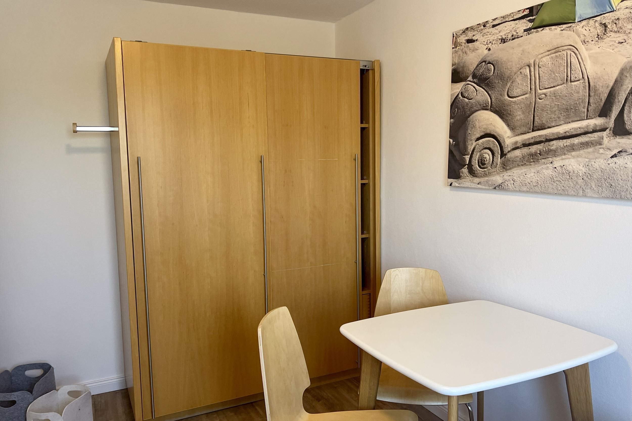 A room with a wooden wardrobe, a white table, and a chair.