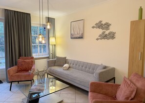 Un salon comprenant un canapé, deux fauteuils, une table basse en verre et une fenêtre avec des rideaux.