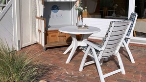 An outdoor seating area with a white table and chairs, a wooden cabinet with a Luna logo, and a glass door. - Luna34 (Room category) - Luna IV (Sankt Peter-Ording)