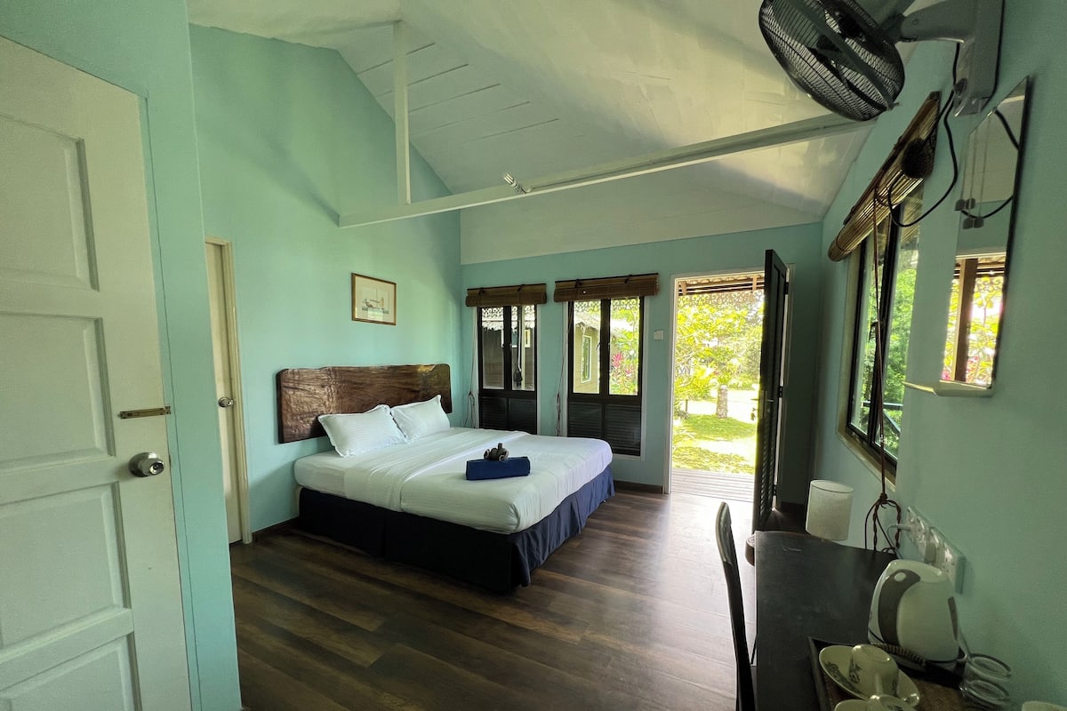 Ein Schlafzimmer mit Bett, Schreibtisch, Stuhl, Ventilator und Fenster mit Blick auf Grünflächen.