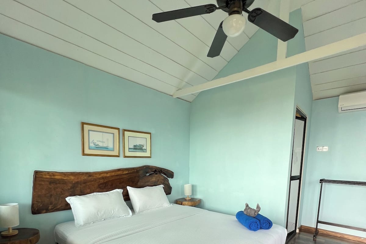 Ein ordentlich bezogenes Bett mit dunkelblauer Bettdecke, einem Deckenventilator und zwei gerahmten Bildern an der Wand.