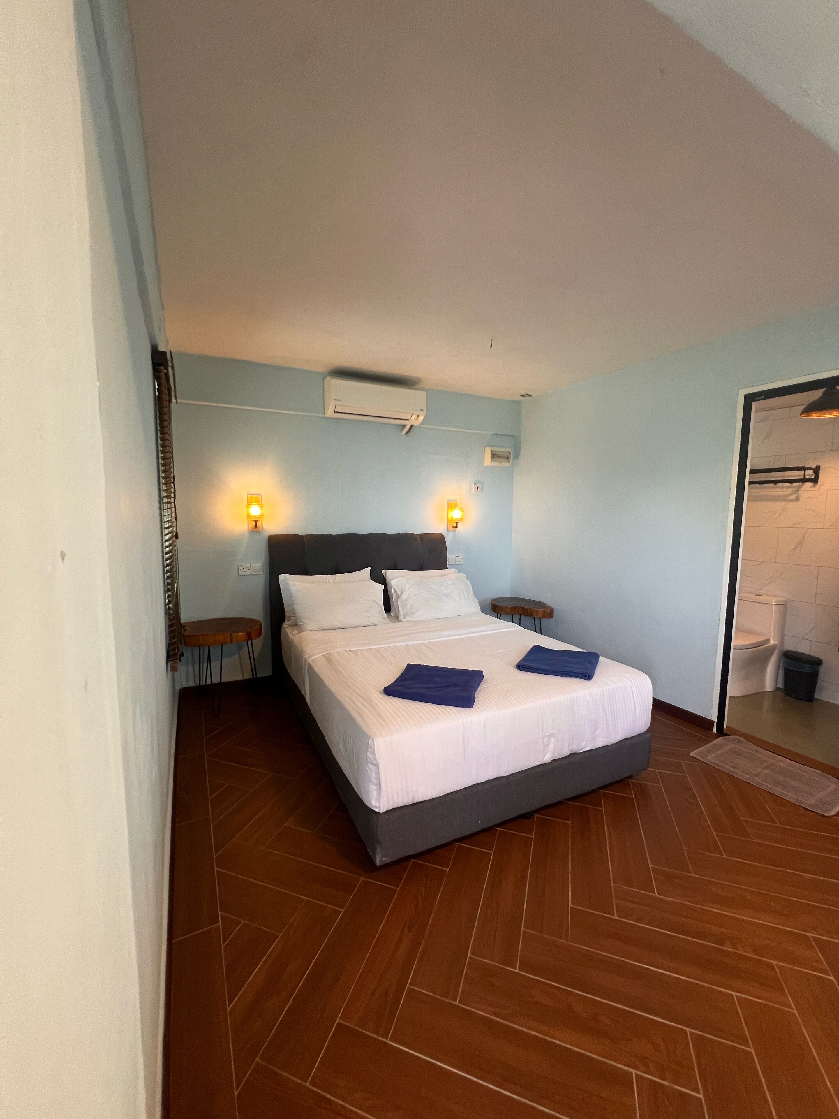 A bedroom with a bed, two bedside tables, a wall-mounted air conditioner, and a bathroom visible through an open door.
