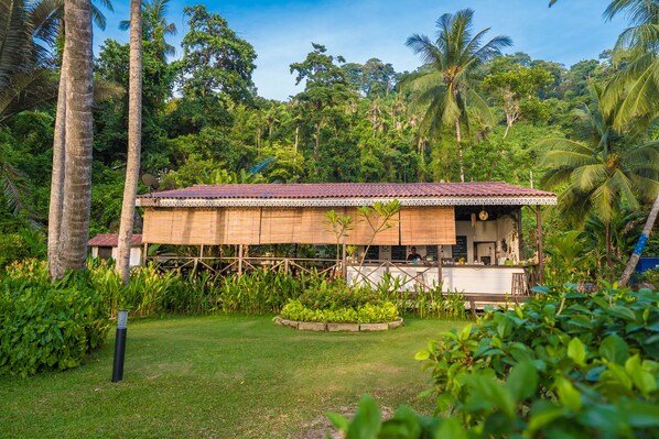 A wooden structure with a tiled roof, surrounded by lush greenery and palm trees. - 1511 Coconut Grove (Tioman Island)