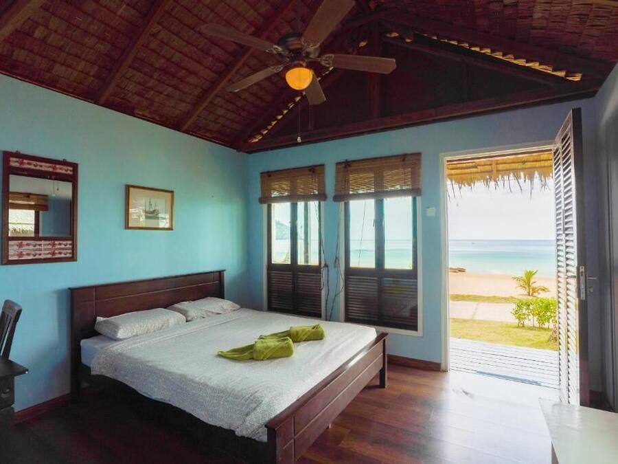 Een slaapkamer met een houten bed, een plafondventilator en uitzicht op het strand.