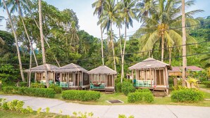A row of thatched-roof huts with green benches, surrounded by lush greenery and palm trees. - 1511 Coconut Grove (Tioman Island)