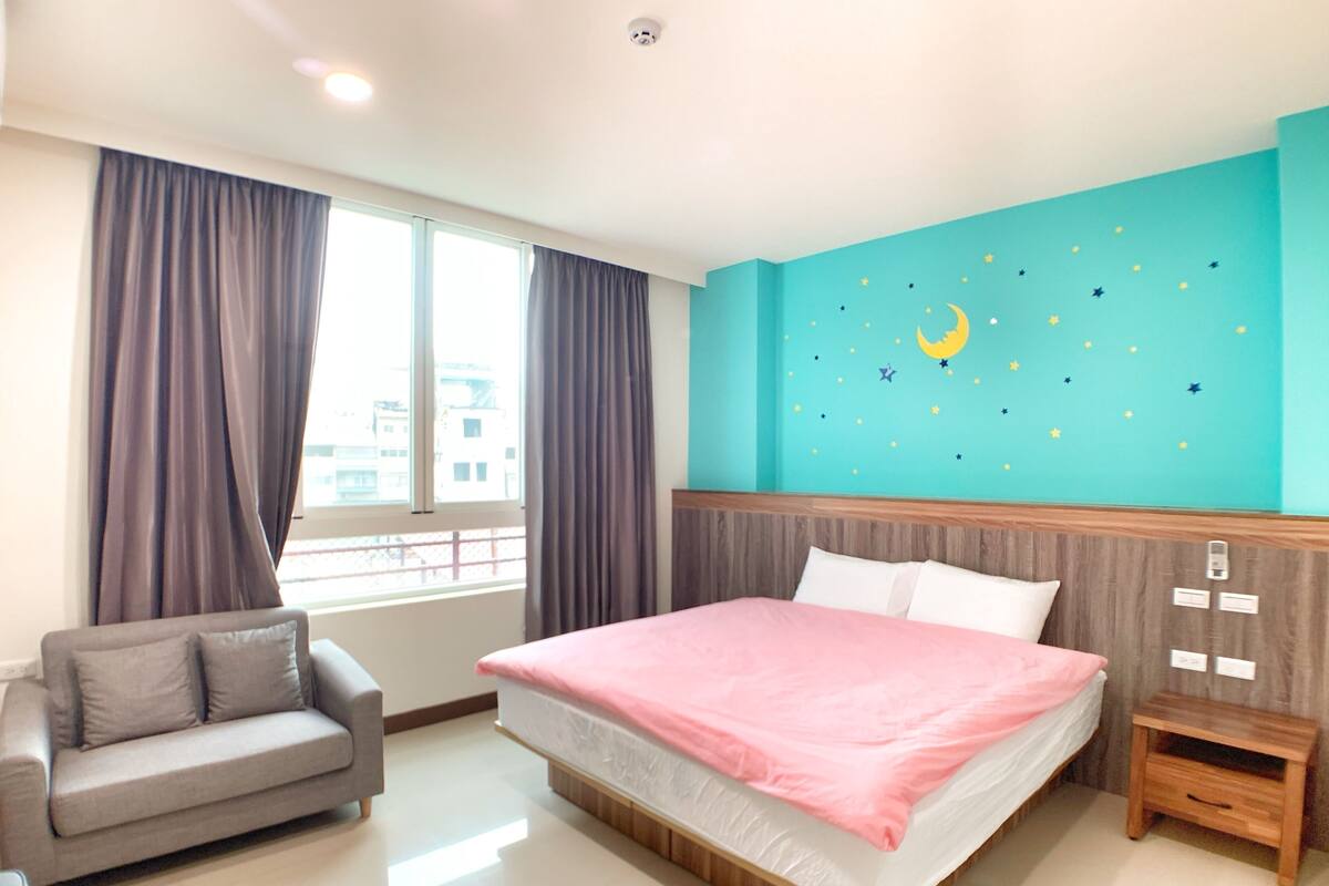 A bedroom with a bed, a grey sofa, a nightstand, a window with curtains, and a wall with star and moon decals.