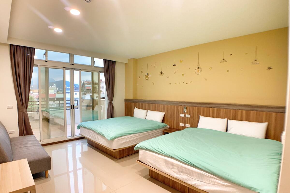 A hotel room with two beds, a sofa, a sliding glass door to the balcony, and a wall with hanging lights and decorative elements.
