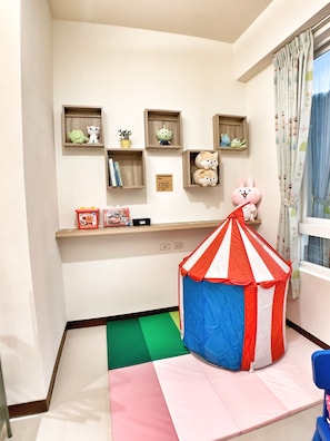 Un área de juegos infantiles con una pequeña tienda de campaña, estantes con juguetes y libros, y una ventana con cortinas.