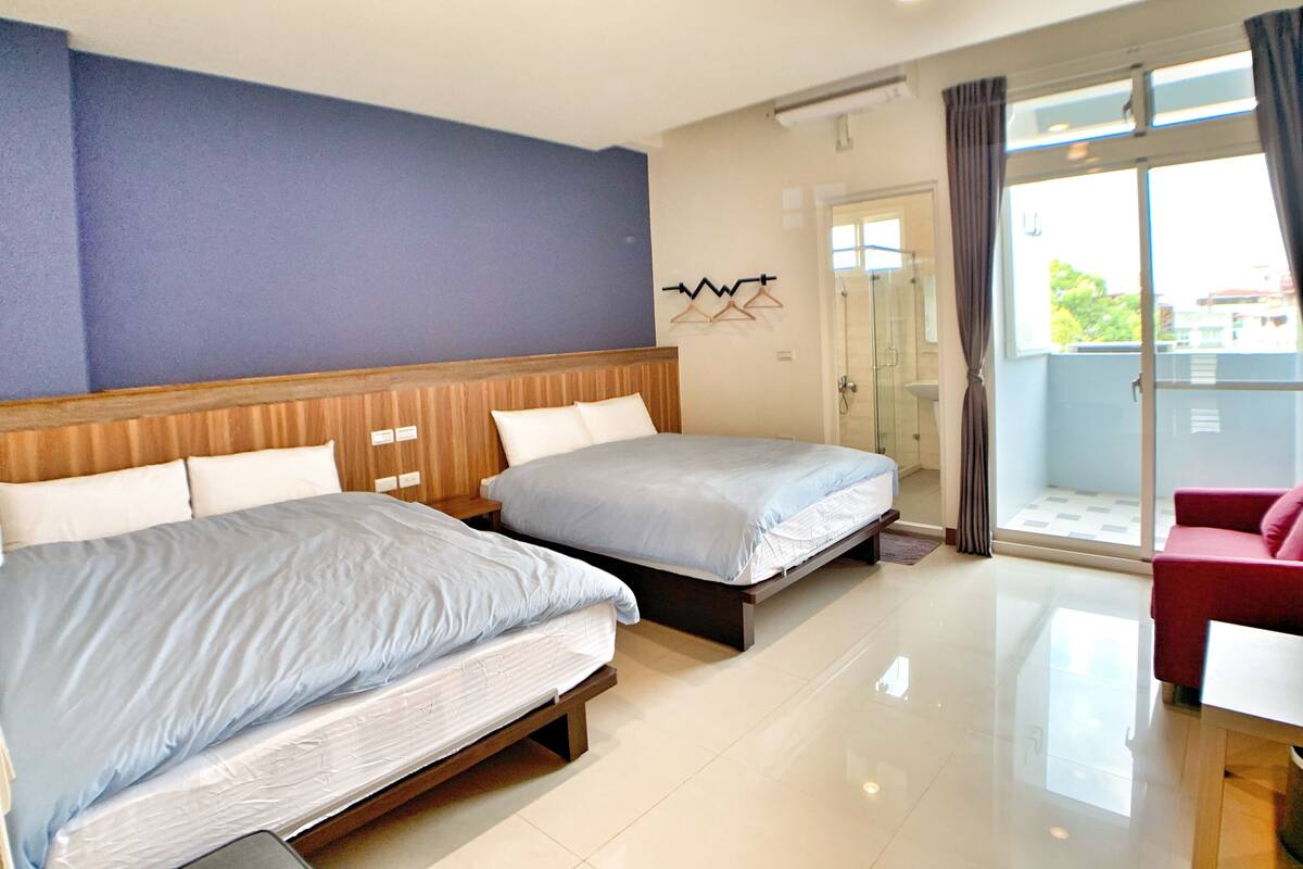 A hotel room with two beds, a balcony with a red chair, and a shower area.