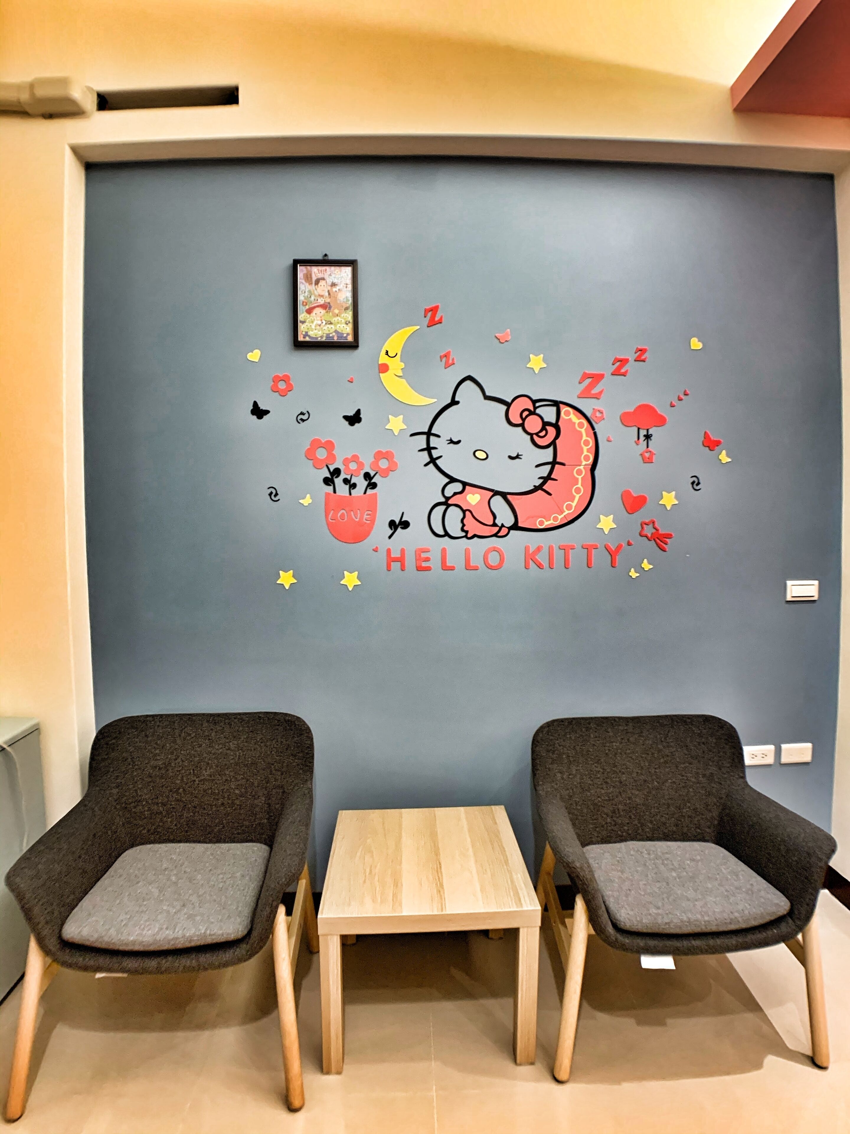 A room with two grey armchairs, a small wooden table, and a wall decorated with a Hello Kitty mural.