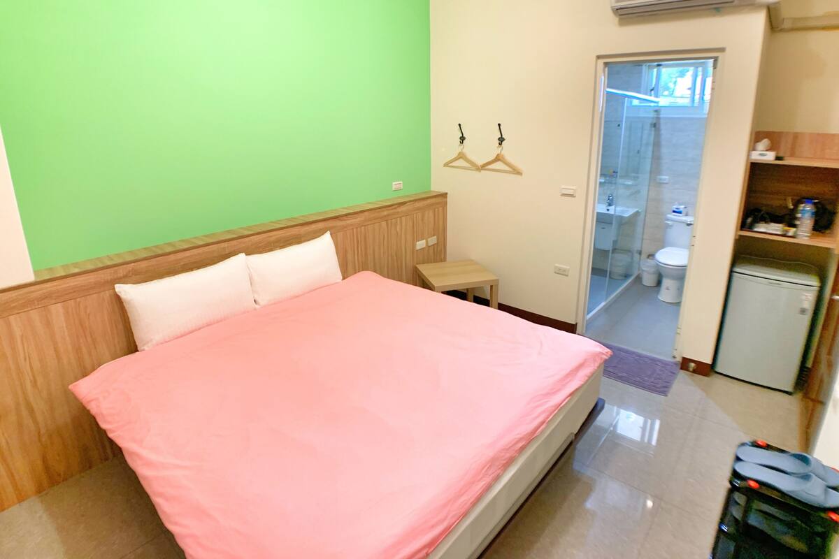 A hotel room with a bed, a small table, a wardrobe, a mirror, and a bathroom visible through a glass door.