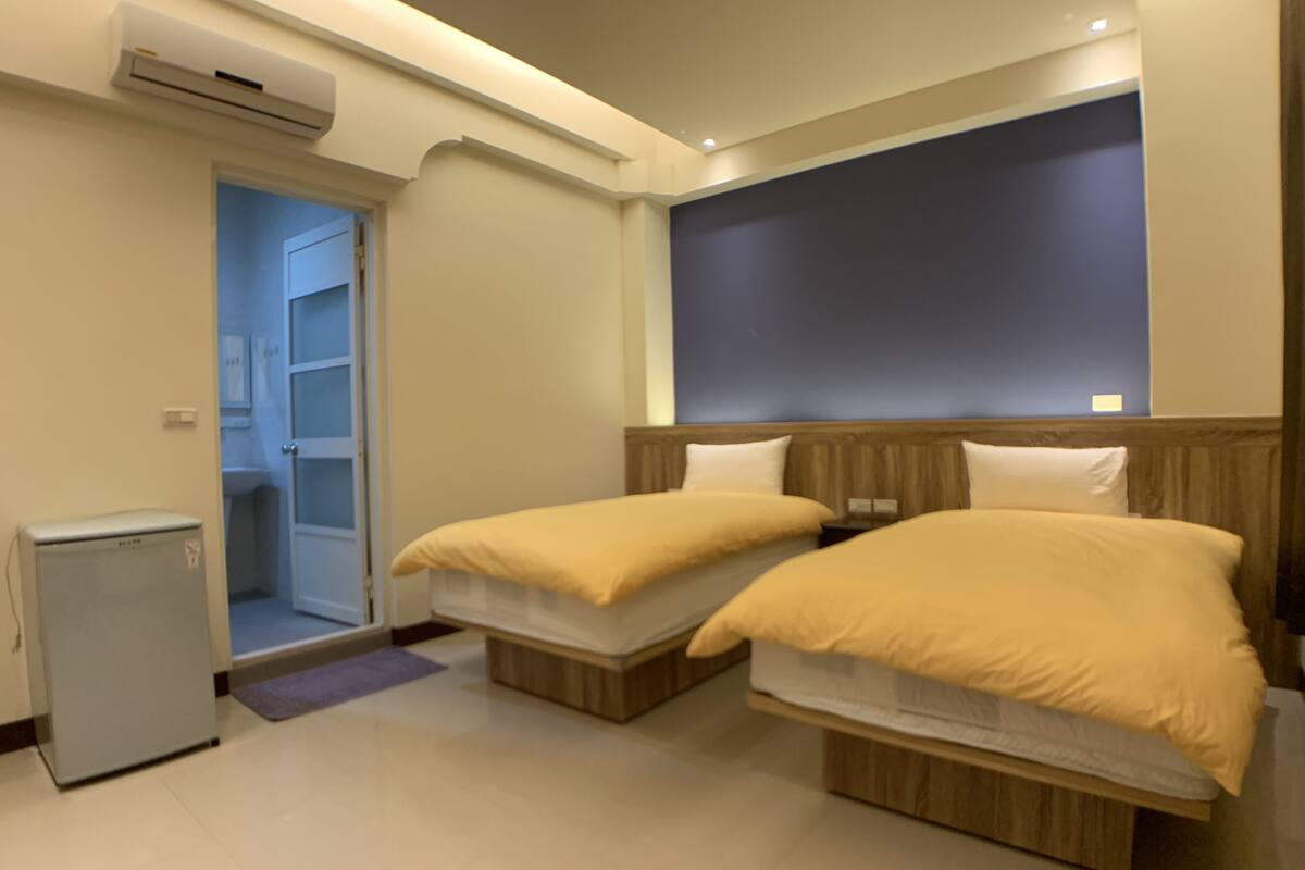 A hotel room with two beds, a small refrigerator, and an air conditioning unit.