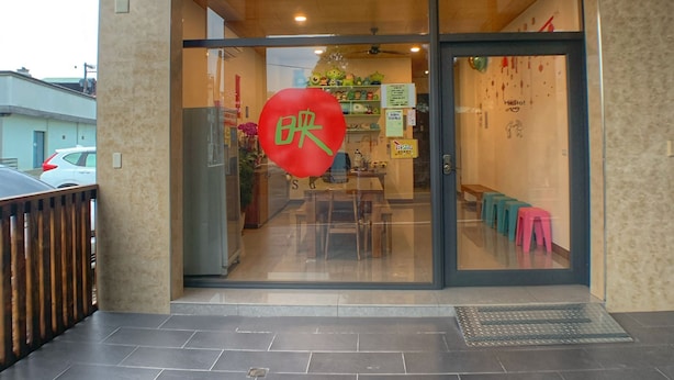 Entrada de un restaurante con puerta de vidrio, un letrero rojo con caracteres chinos y un área de mesas visible a través del vidrio.