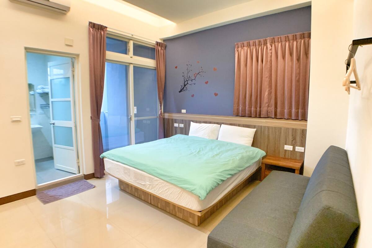 A modern bedroom with a bed, a grey sofa, and a bathroom visible through a glass door.