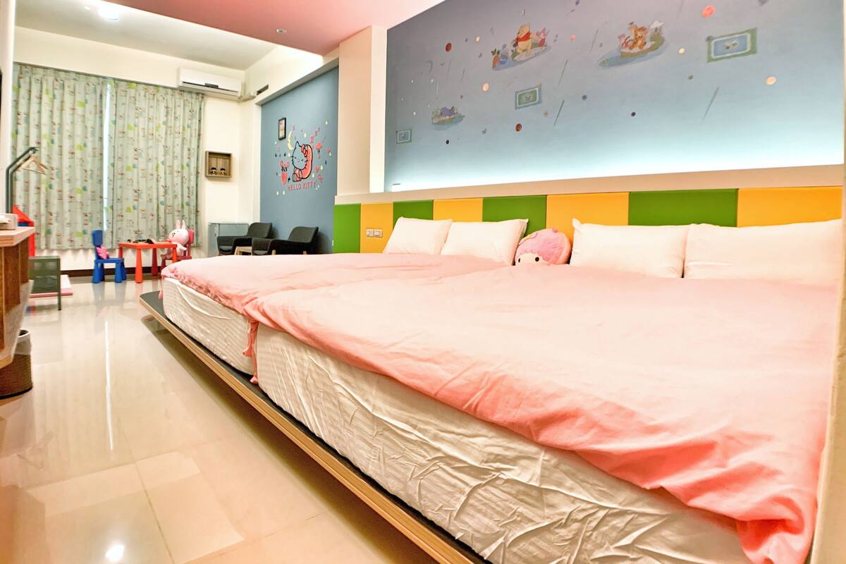 A hotel room with a large bed, a desk, a chair, a small table, and a wall with a Hello Kitty mural.