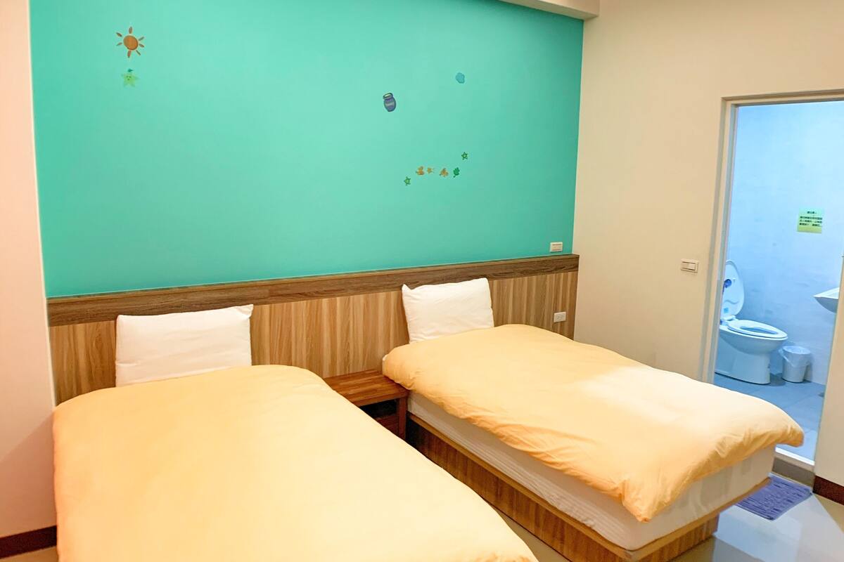 A hotel room with two beds, a teal wall, and a visible bathroom door.