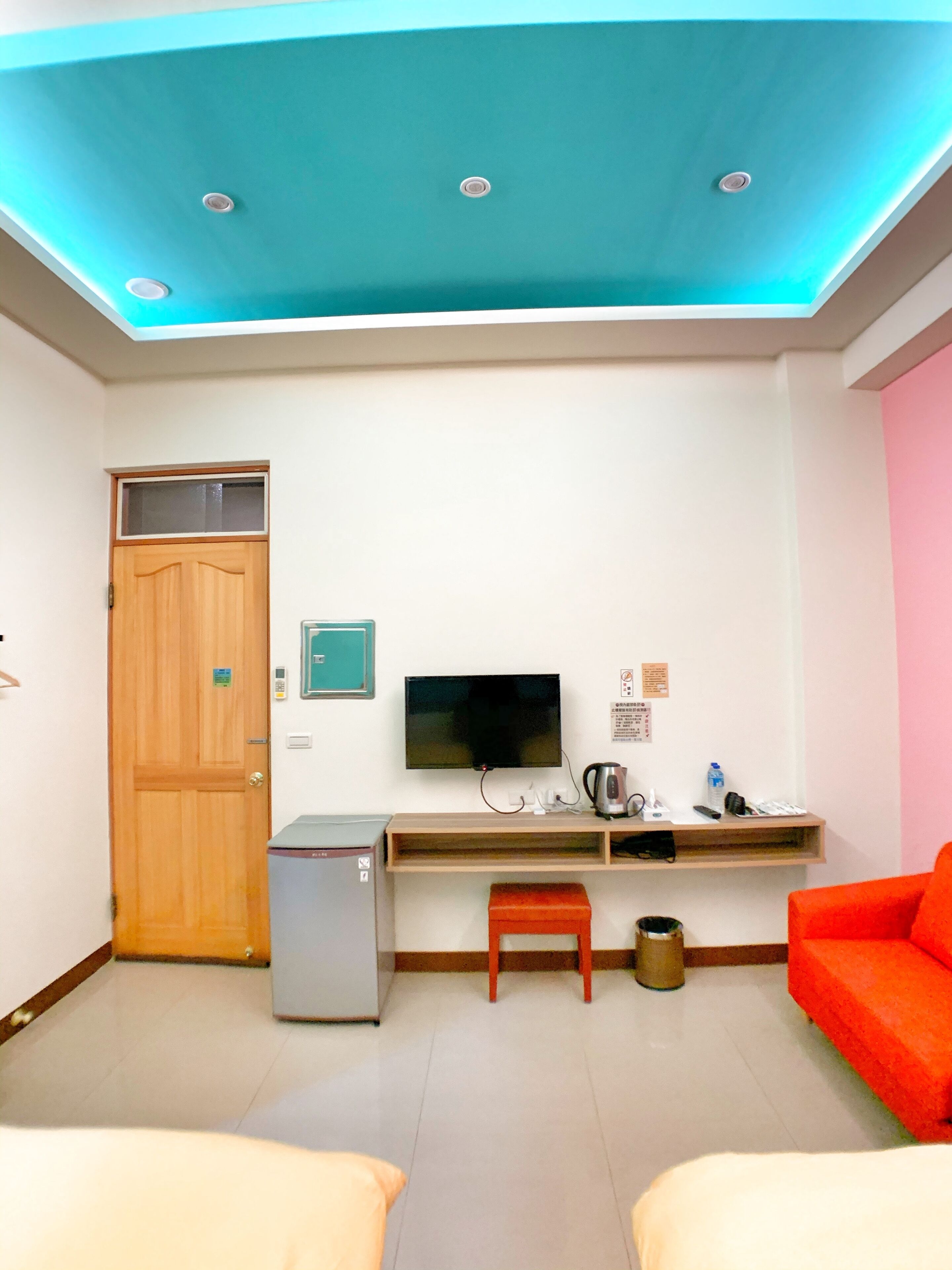 A room with a wooden door, a TV mounted on the wall, a small desk with a kettle, a red chair, and a bed.