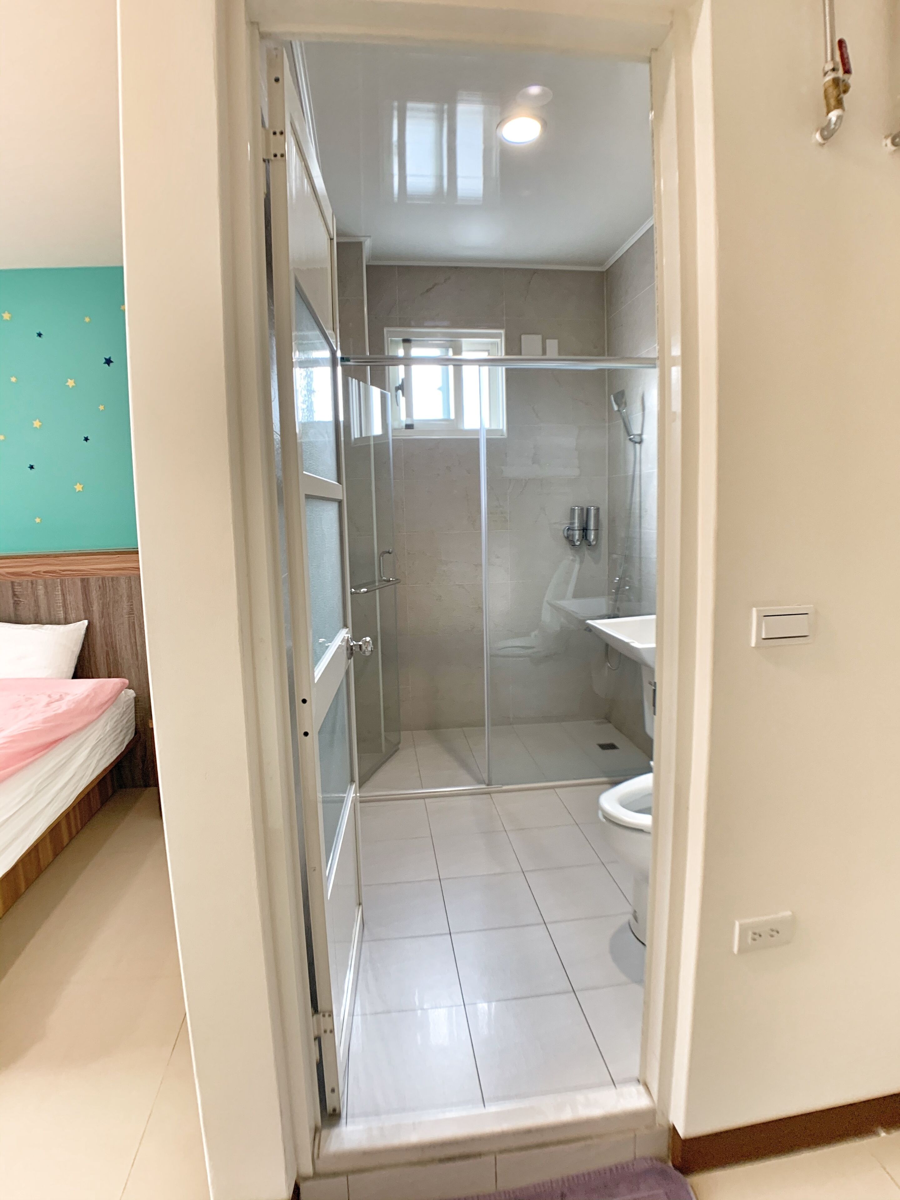 A room with a glass-enclosed shower, a bed with a pink and white duvet, and a teal wall with star decorations.