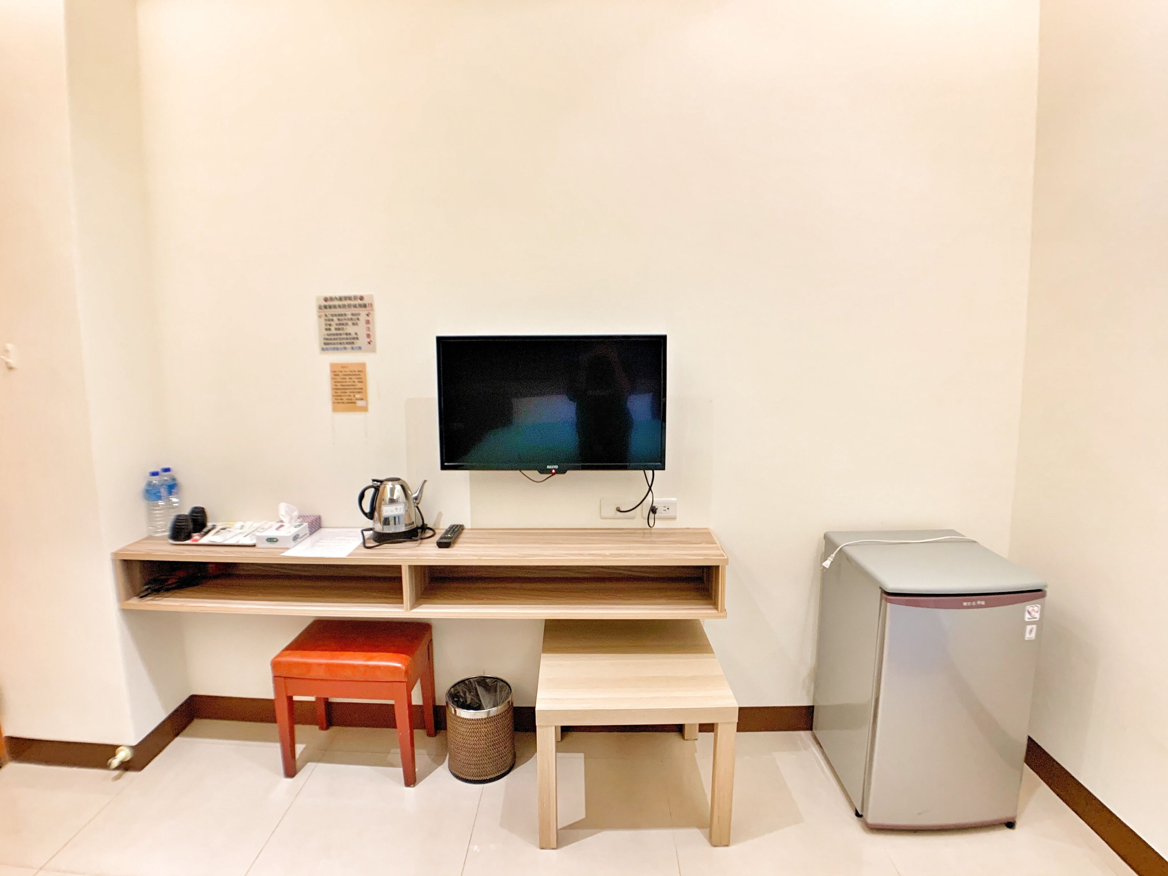 A small room with a desk, a television, a refrigerator, a trash can, and a small stool.