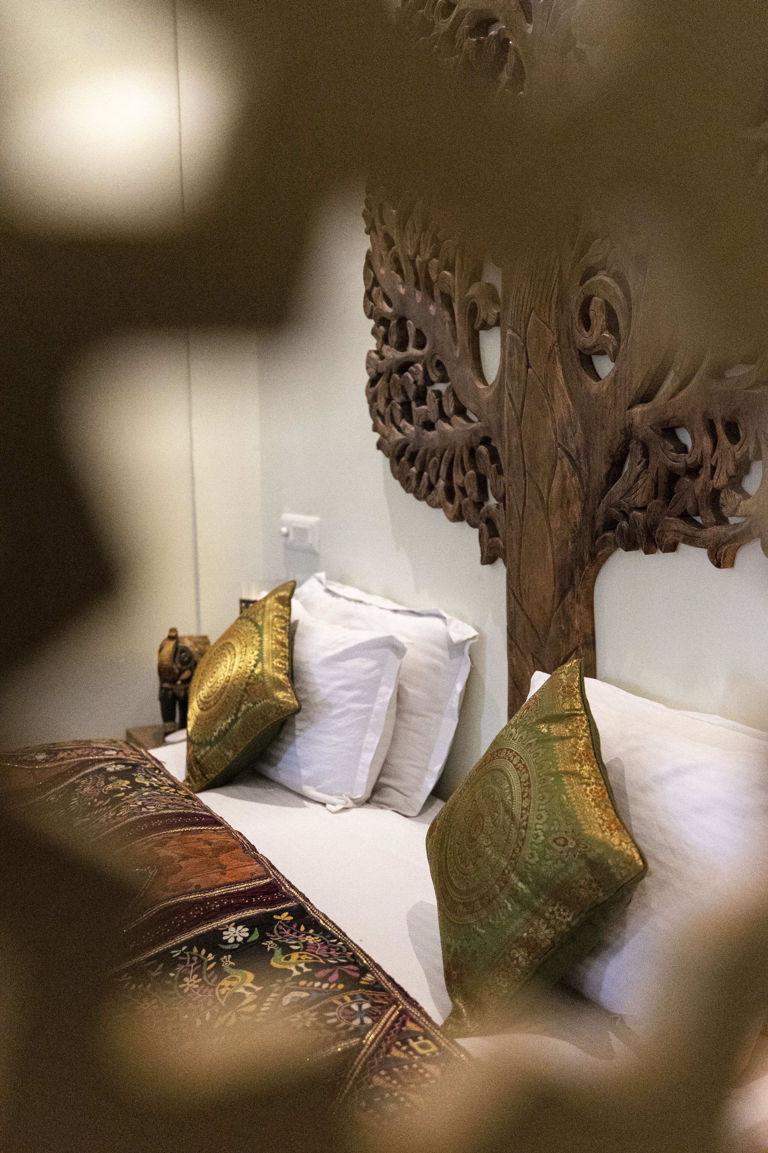 A bed with white and gold pillows, a patterned throw, and a carved wooden headboard.