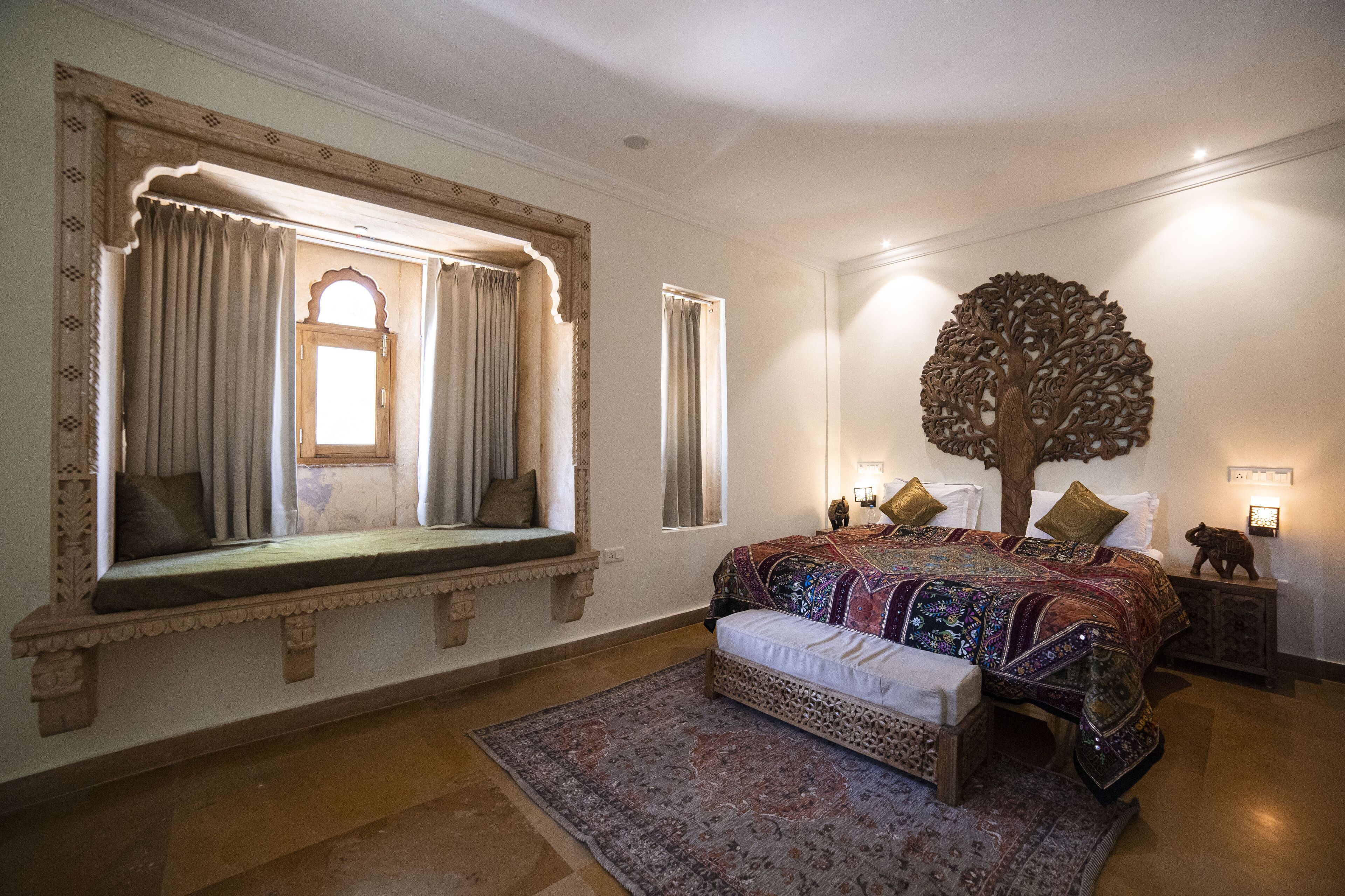 A bedroom with a large bed, a bench with a view, a tree wall decoration, and a patterned rug.