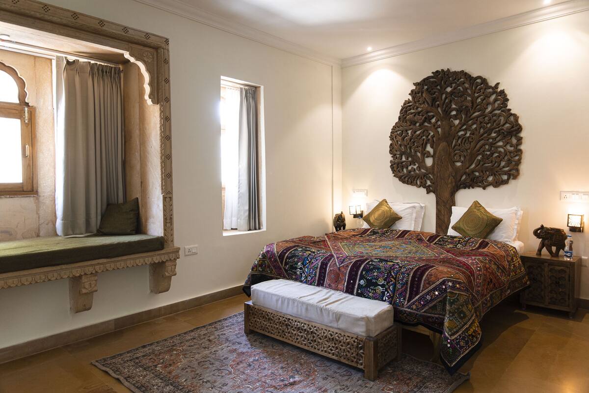 Een slaapkamer met een groot bed, een bankje, decoratieve wandkunst met een boom en een raam met gordijnen.