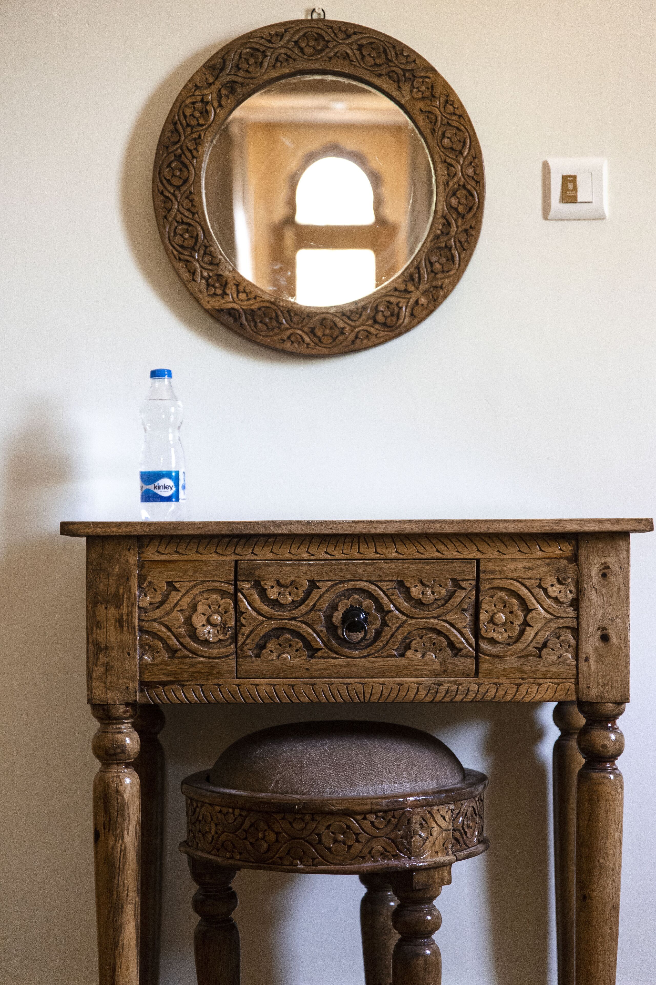 A wooden console table with intricate carvings and a round mirror above it. A bottle is placed on the table.