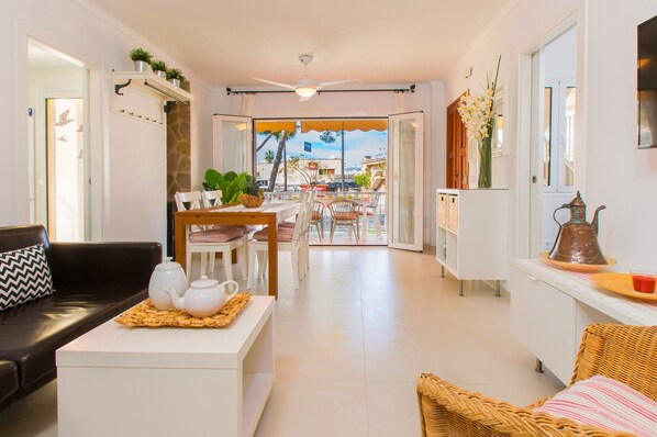 A modern living room with a sofa, a dining area with chairs, a kitchen area with a counter, and a view through a glass door to the outside.